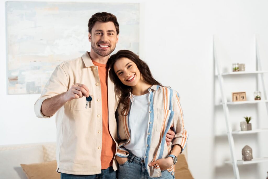 Jovem casal sorrindo para a câmera enquanto segurava chaves de nova casa na sala.