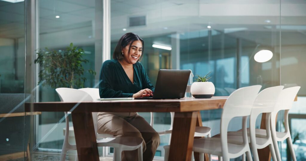 Mulher jovem em escritório com ambiente bem iluminado de frente para um notebook aberto e sorridente.