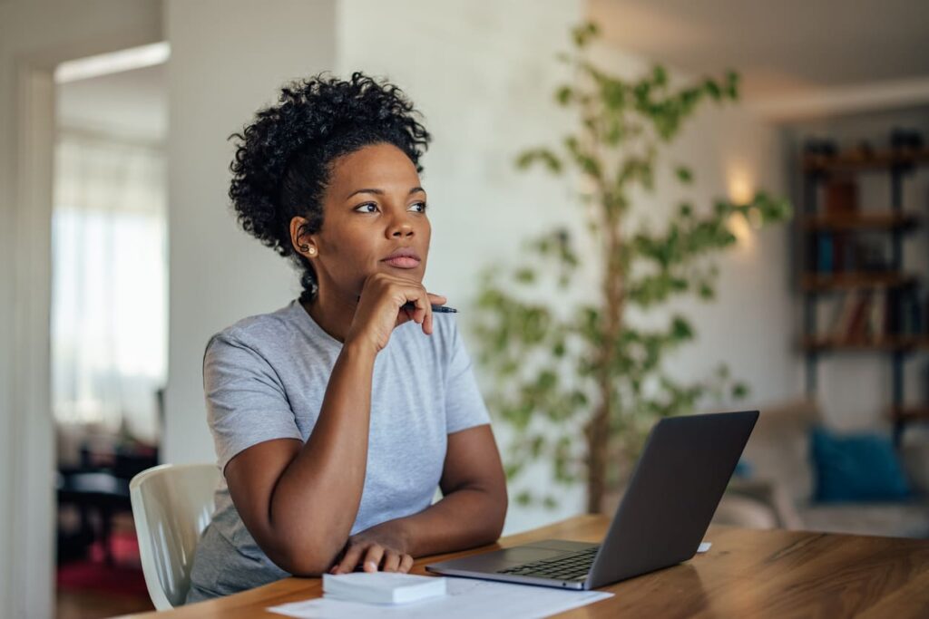 Jovem sentada de frente pra mesa com seu notebook aberto e expressão de pensamento com a mão no queixo.