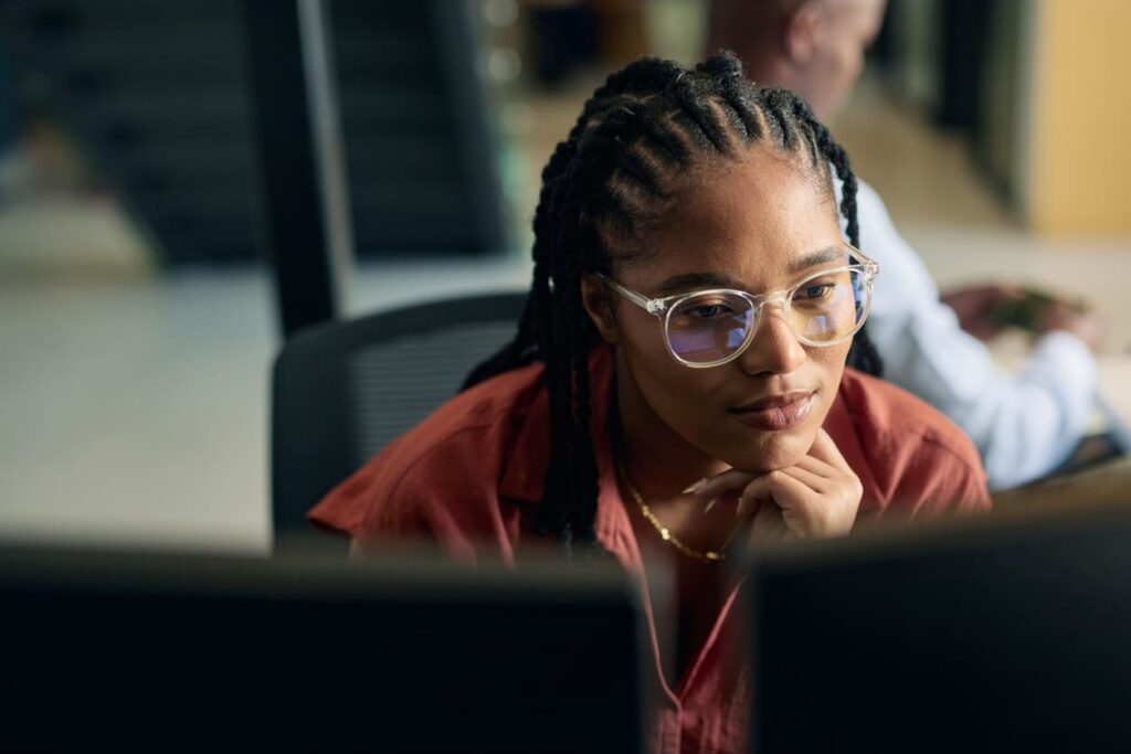Mulher jovem em escritório de frente para o computador com expressão pensativa.