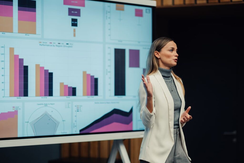 Mulher executiva, usando um terno branco, dando uma palestra com slides que possuem infográficos ao fundo.
