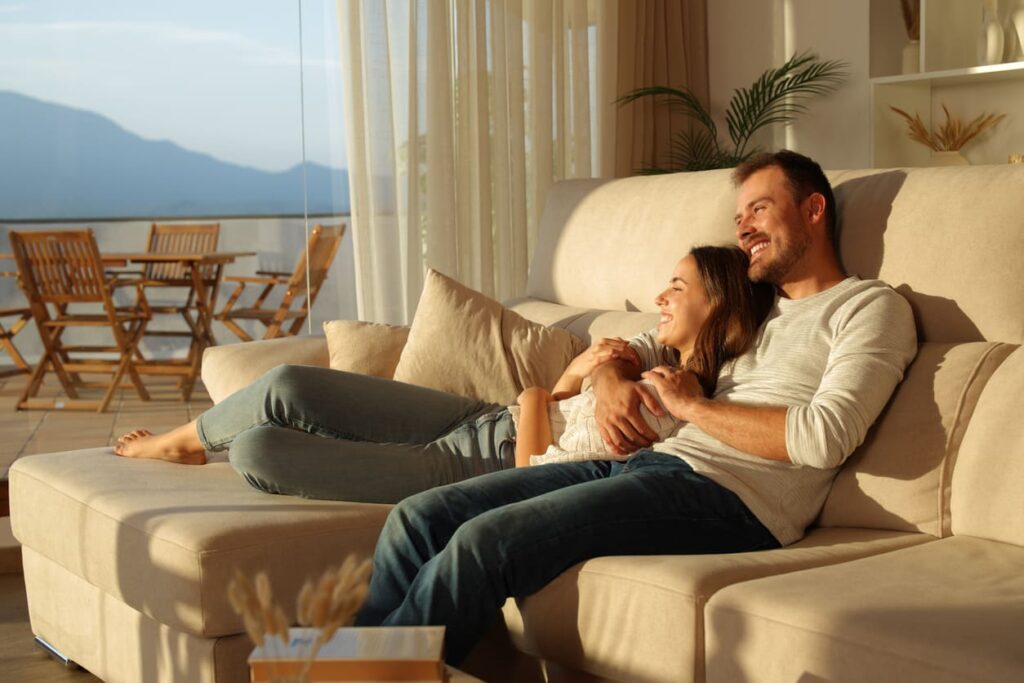 Casal feliz ao pôr do sol em casa, sentados no sofá, olhando através de uma janela.