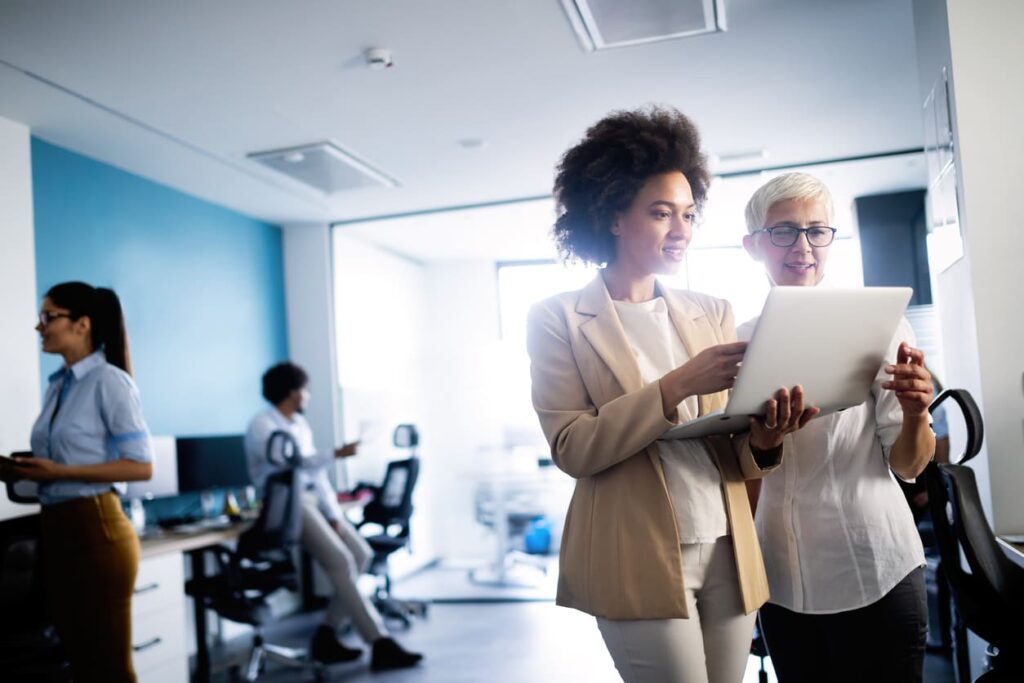 Duas mulheres em ambiente corporativo, com roupas formais, analisando algo em um notebook branco.