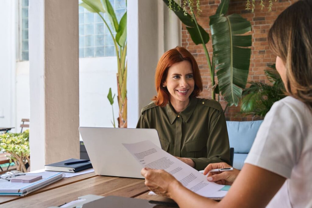 Duas mulheres em sala de escritório conversando com papéis e notebook na mesa.