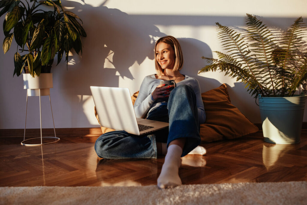 Mulher sentada no chão do apartamento com um notebook nas pernas e recebdno a luz do sol.