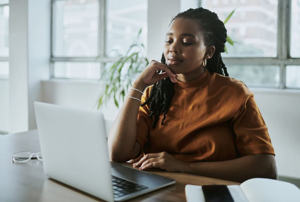 Mulher em frente ao computador em um ambiente claro com a mão no queixo pensativa.
