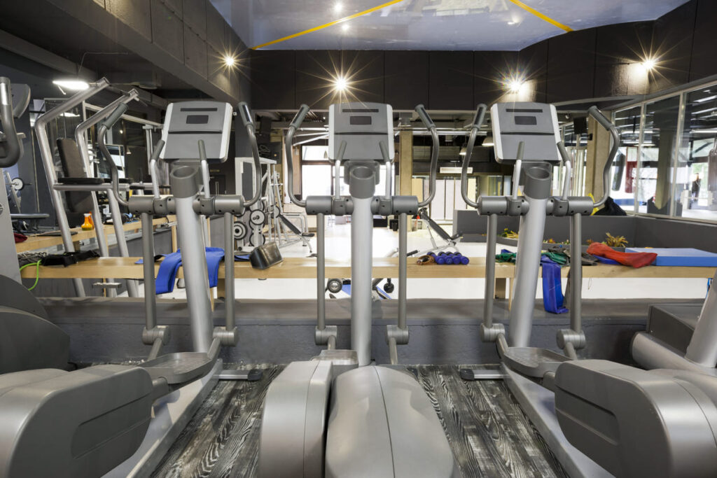 Foto de esteiras da cor cinza em academia com espelho ao fundo e ambiente iluminado.