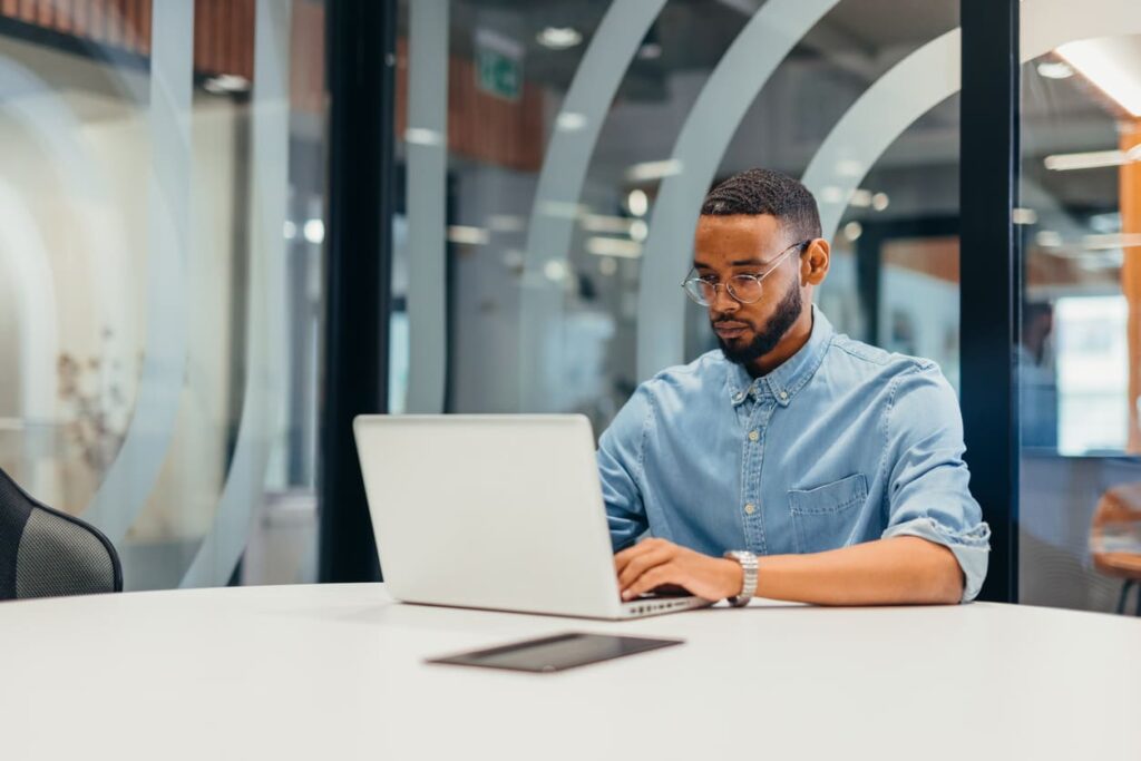 Um homem está sentado em uma sala de escritório bem iluminada com um notebook aberto na sua frente.